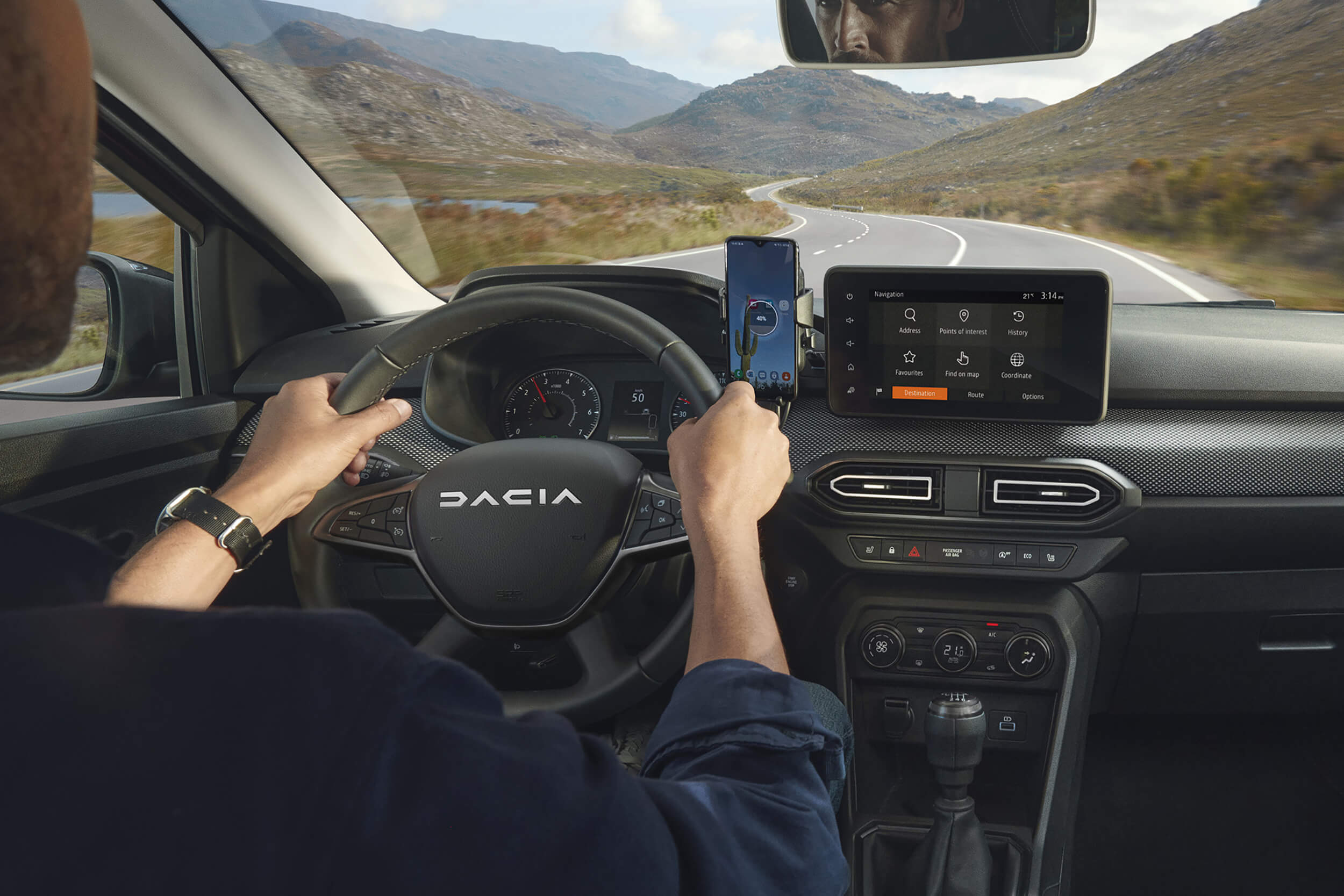 Vyras vairuoja Dacia Jogger, matomas vairas, prietaisų skydelis, centrinis ekranas ir konsolė, išmanusis telefonas laikiklyje