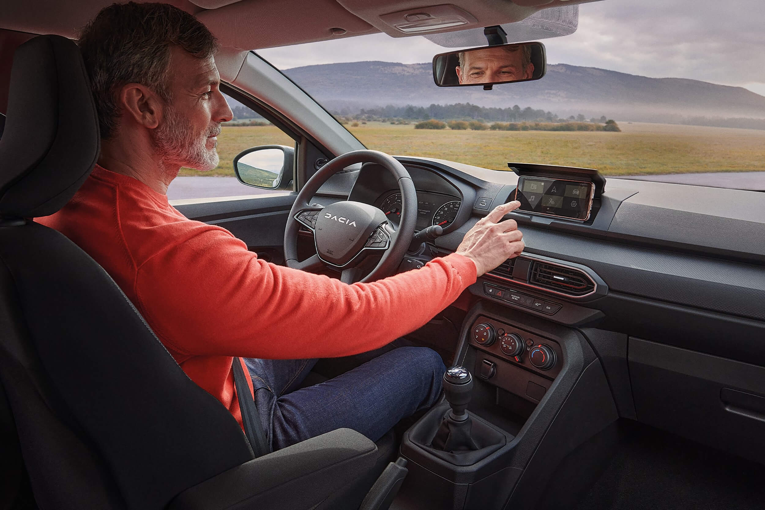Vyras sėdi prie Dacia Sandero Stepway vairo ir valdo MEDIA CONTROL sistemą laikyklyje pritvirtintame telefone