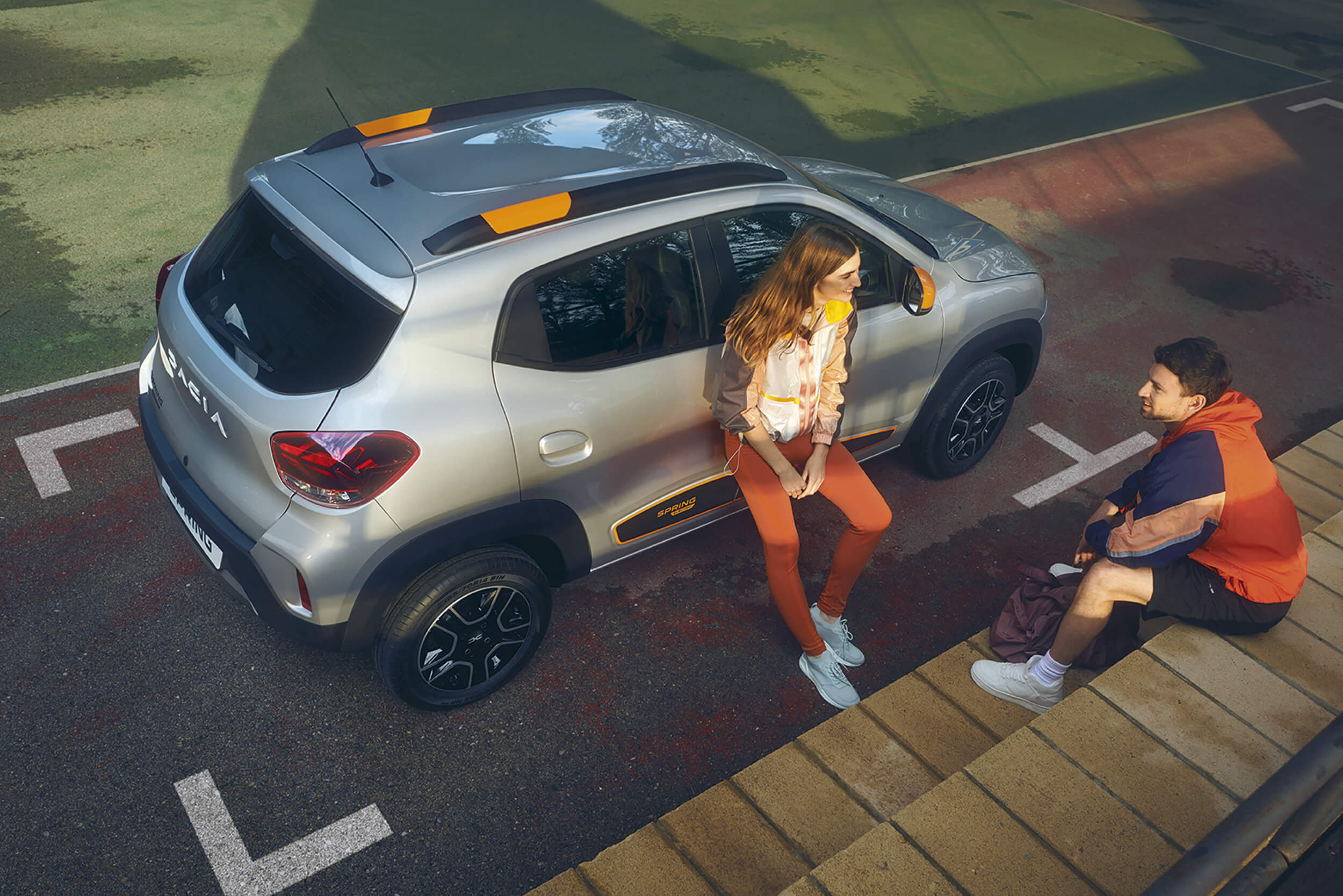 Elektromobilis Dacia Spring pastatytas stovėjimo vieta, šalia stovi į automobilį atsirėmusi mergina